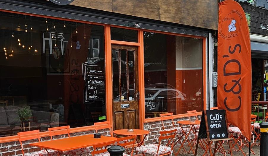 Exterior of coffee shop with orange trimmed windows, open sign, orange metal tables and chairs with cushions.