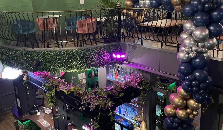 Interior of Nanouk. A stylish bar interior with an upper seating area featuring pink and teal chairs, green walls, and hanging plants. A large balloon