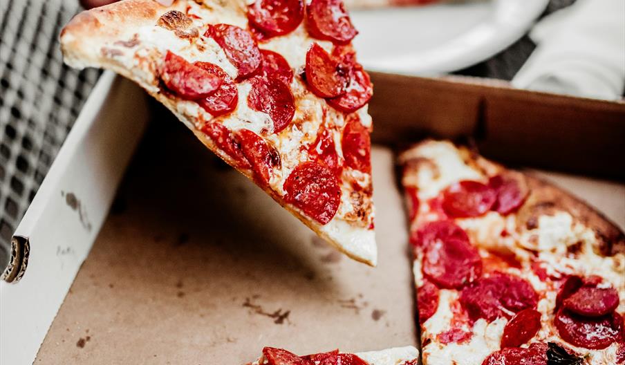 A pepperoni pizza in a wooden box, with someone taking a slice. In the background is a white plate with another slice already served.