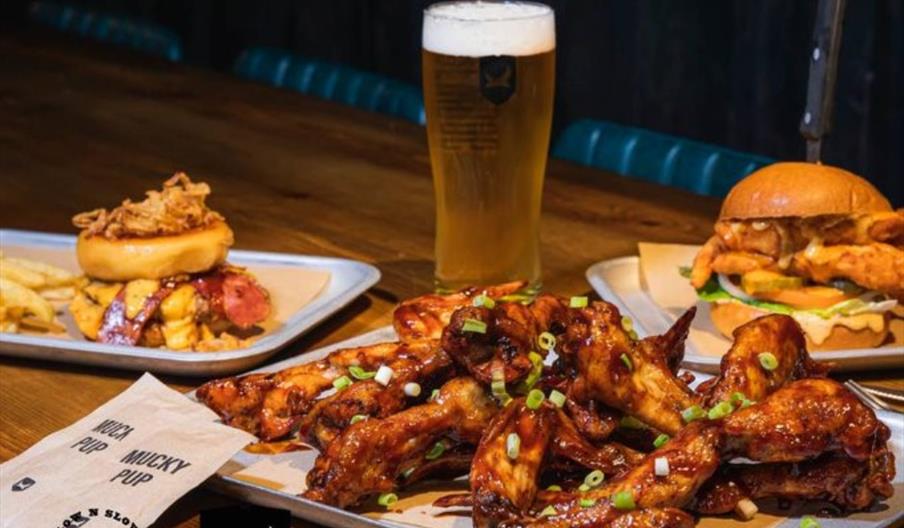 Chicken wings on a white plate with burger and a pint of beer on a wooden table.