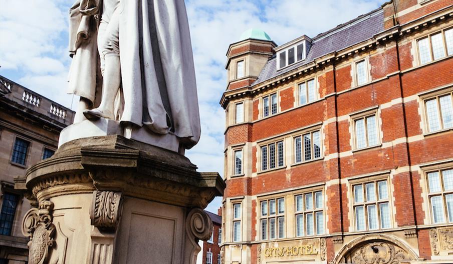 Red brick building with a gold sign that reads City Hotel, with a statue in the foreground under a cloudy sky.