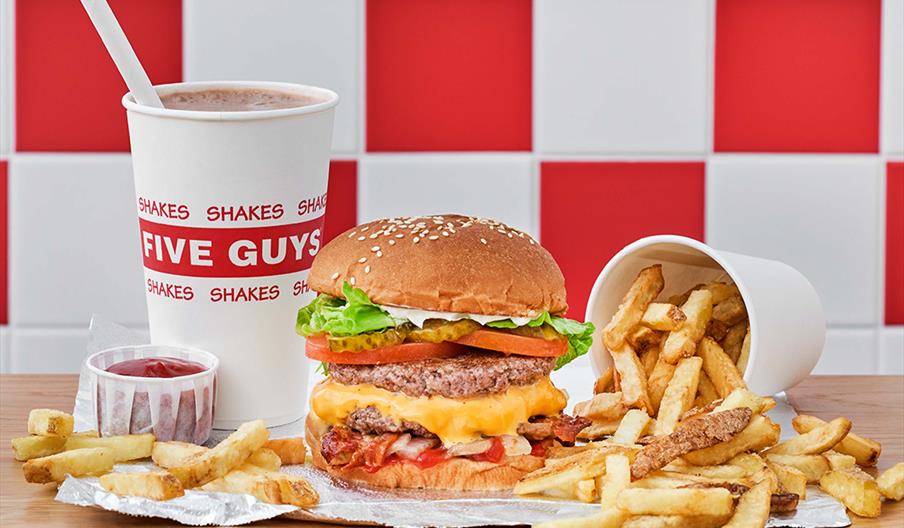 A Five Guys meal featuring a double cheeseburger with lettuce, tomato, and bacon, accompanied by a cup of fries spilling onto the table, a ketchup con