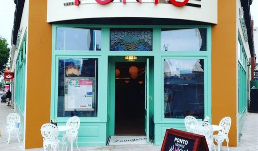 Exterior of "PONTO" cafe with mint-green framed windows and doors, mustard-yellow accents, and outdoor seating with white metal chairs and tables. A s