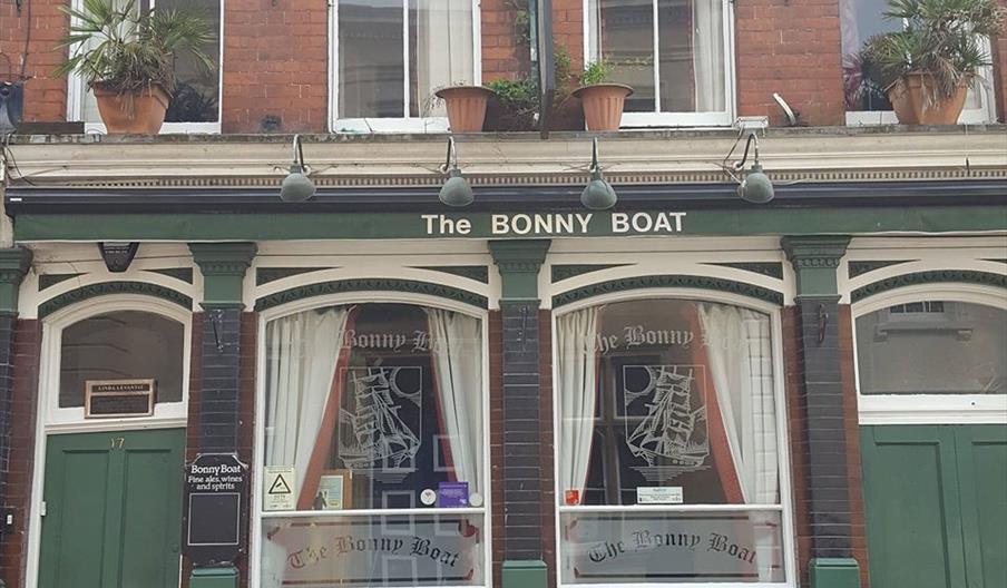 Street view of "The Bonny Boat" pub, featuring large sash windows with ship designs, green trim, decorative yellow tiles below the windows, and potted