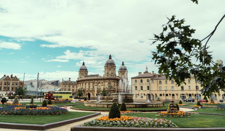 A fountain surrounded by flowerbeds and grassy areas, with the ornate sandstone building of the Hull Maritime Museum behind it, complete with its dome