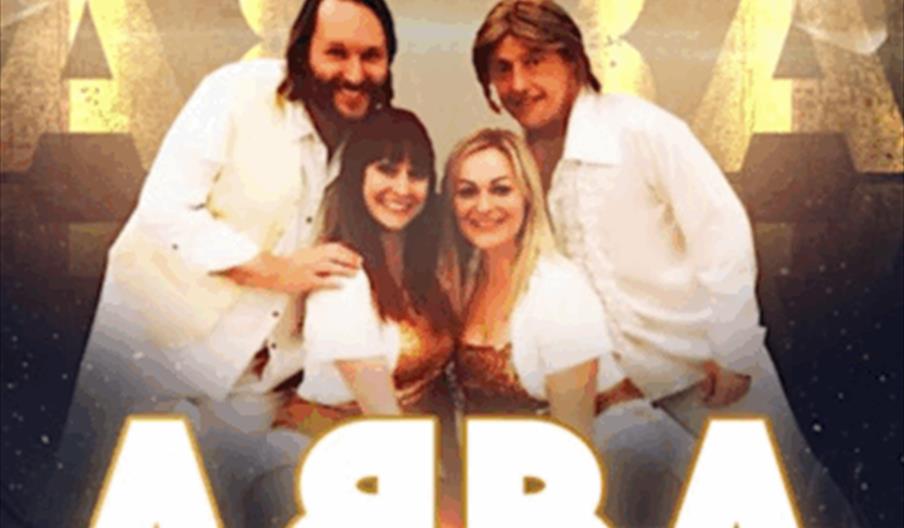 A group of four people dressed in white pose and smile in front of a gold-lit backdrop with the large word "ABBA" and spotlights; the text "ABBA UK" a