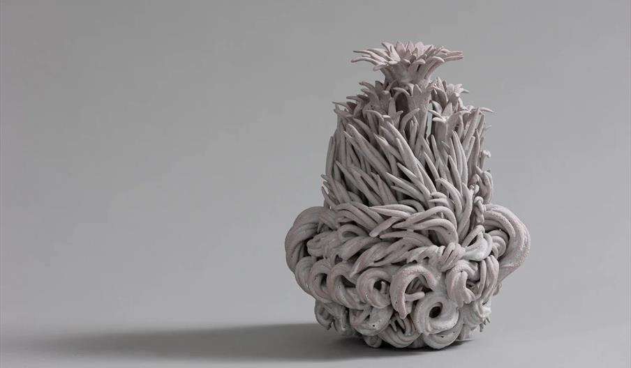 A sculpted ceramic vase with an intricate, twisting design that resembles clusters of curled tendrils and spiky, plant-like forms, set against a plain