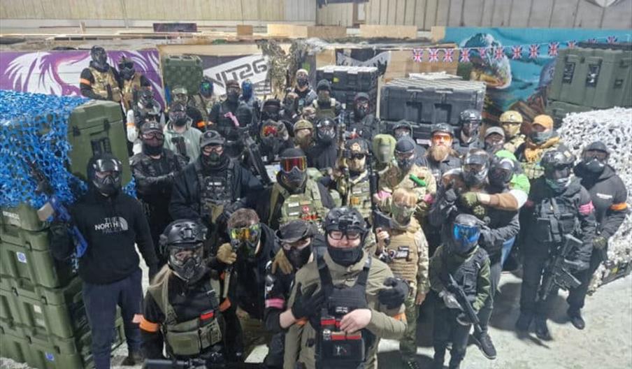 A large group of people in tactical gear and face protection pose for a photo inside an indoor airsoft arena, surrounded by stacked crates and camoufl