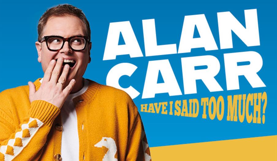 Alan Carr in a yellow sweater with a white design covers his mouth while smiling, standing in front of large text that reads "ALAN CARR" and "HAVE I S