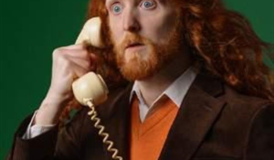 A person with long red hair and a beard, wearing a brown suit and orange sweater, holds a vintage cream-colored telephone to their ear and looks surpr