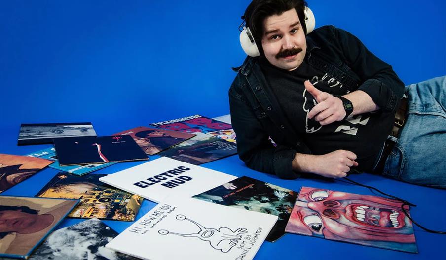 A man with headphones and a mustache is lying on his side on a blue floor, surrounded by vinyl record covers, and pointing toward the camera.
