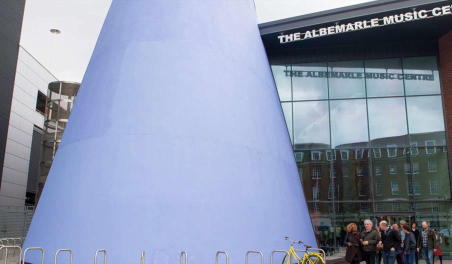 Large blue conical sculpture in front of The Albemarle Music Centre, with a group of people gathered near the entrance and a yellow bicycle parked by