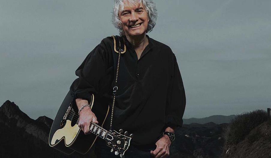 Promotional poster for an Albert Lee concert, showing a smiling man with gray hair holding a guitar and standing on a road with mountains in the backg