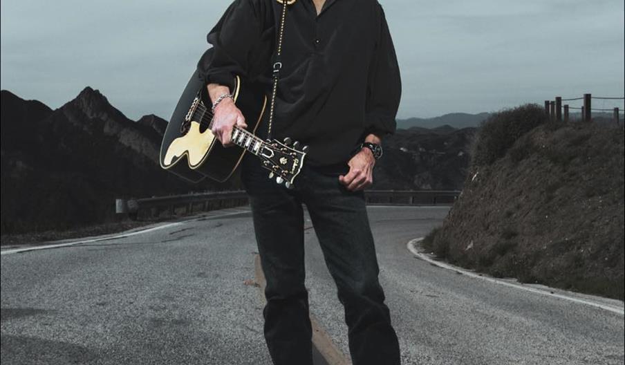 A man with grey hair wearing all black stands smiling whilst holding a guitar in the middle of the road.