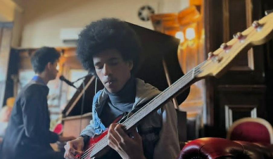 Two young men are playing music in an elegant room; one is in the foreground playing an electric bass guitar, while the other is seated at a grand pia
