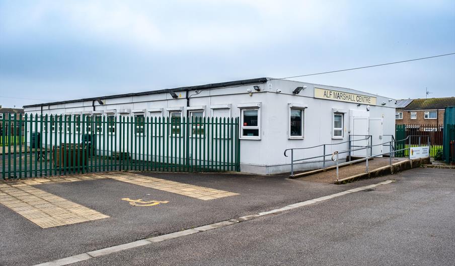 A single-story white modular building with a sign reading "ALF MARSHALL CENTRE," surrounded by a tall green fence, features a wheelchair-accessible ra