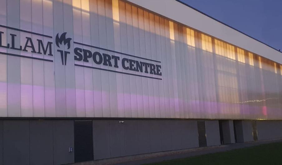 Exterior view of the Allam Sport Centre building at dusk, with illuminated panels casting a warm glow and the centre’s name displayed prominently on t