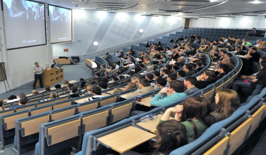 A large, modern lecture hall filled with students seated in tiered rows, listening to a person giving a presentation at the front near a podium, with