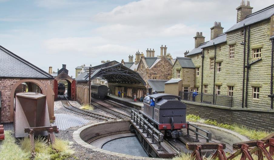 A detailed model railway scene featuring a small train station with covered platforms, realistic buildings, and a turntable with a model train. The sc