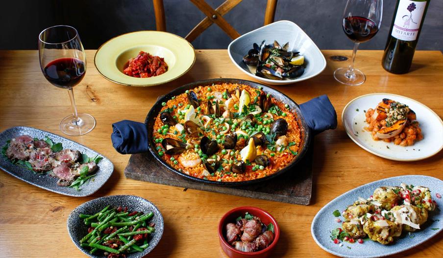 A table set with a large pan of seafood paella in the center, surrounded by various small plates of appetizers including green beans, sliced meat, she