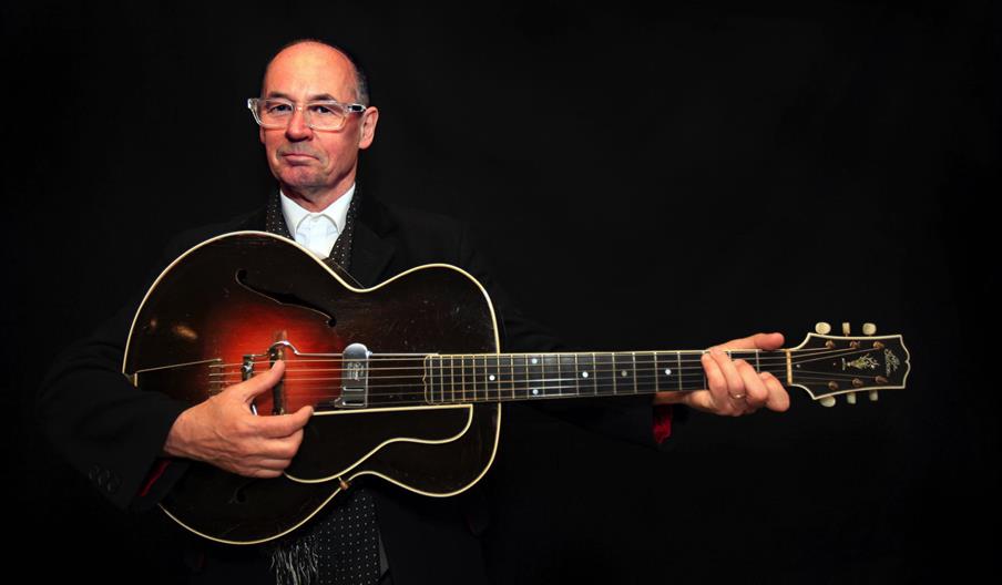 Andy Fairweather Low wearing glasses and formal clothing holds an acoustic guitar, positioned as if ready to play, against a plain black background.