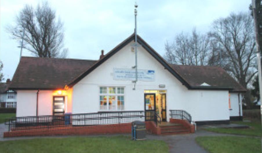 A small white community building, Anlaby Library with a brown shingled roof, wheelchair-accessible ramp, and steps leading to the entrance. There are