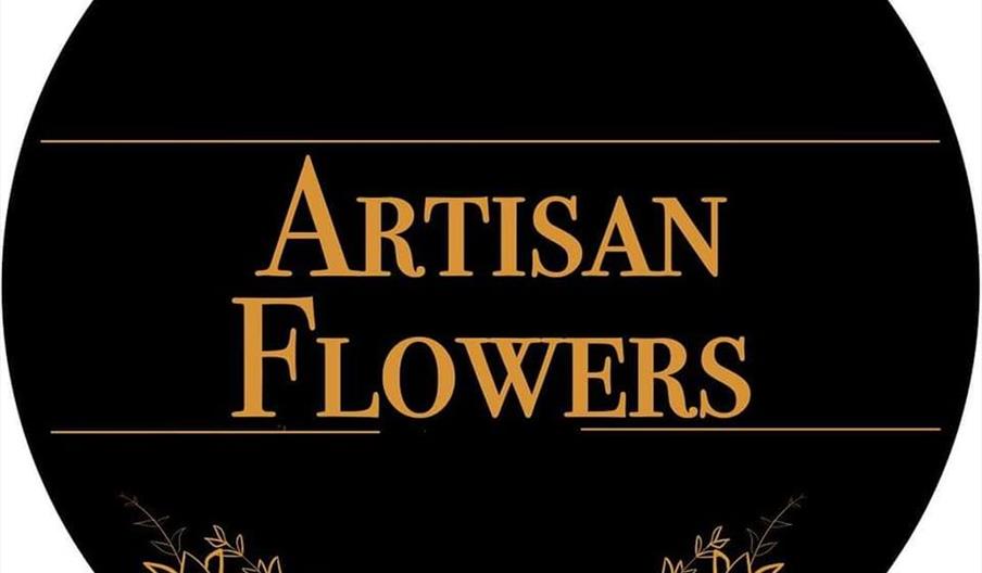 A black circular logo with the words "Artisan Flowers" in gold lettering, framed by gold horizontal lines and accented with a decorative gold leafy br