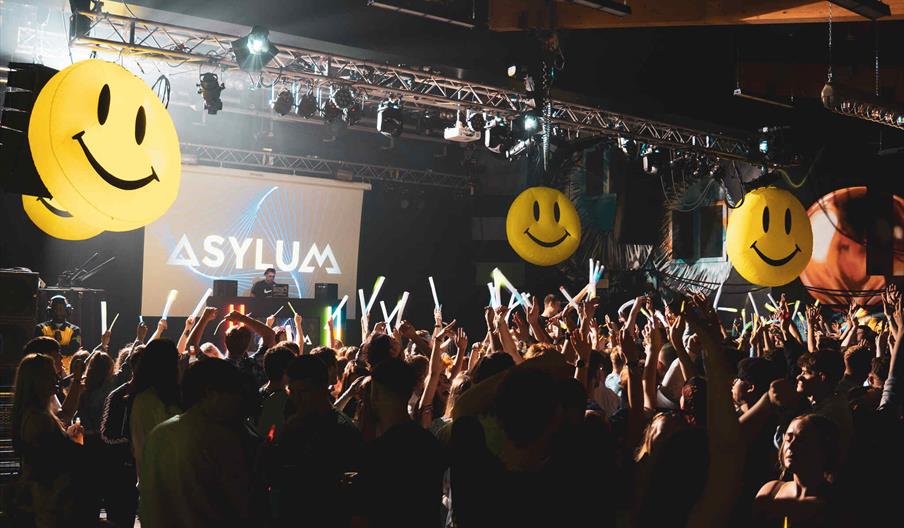 The interior of Asylum nightclub in Hull. The room is packed with clubbers holding glowsticks in the air, whilst a DJ performs on a stage in front of