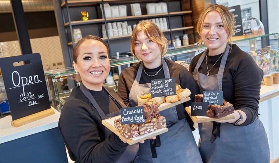 Three smiling café staff members wearing gray aprons stand behind the counter, each holding a tray with baked goods labeled "Crunchie Rocky Road," "Ch