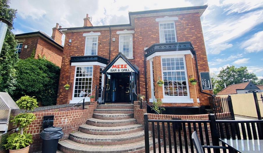 A brick building with large windows and a sign reading "Meze Bar & Grill" above the entrance, accessed by wide curved steps, with outdoor seating and