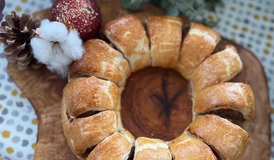 A golden-brown ring-shaped sausage roll is displayed on a rustic wooden serving board, garnished with a pine cone, a red glittery ornament, and a piec
