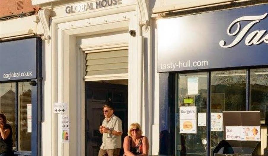 Three people sit and stand outside a storefront called "Tasty". One woman sits behind an ice cream freezer with signs advertising ice cream; a man sta