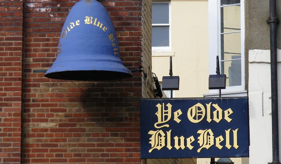 An exterior shot of the alley leading to Ye Olde Blue Bell. On the corner of a brick wall is a blue sign and a blue bell both featuring gold writing t