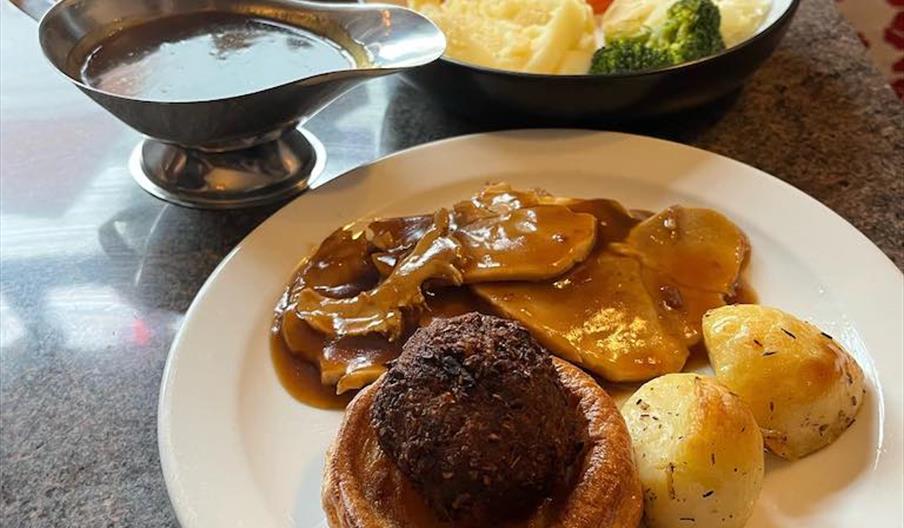 A plate with of Sunday roast with potatoes, yorkshire pudding, beef and stuffing. Next to it sits a gravy boat and a bowl of carrots, cauliflower and
