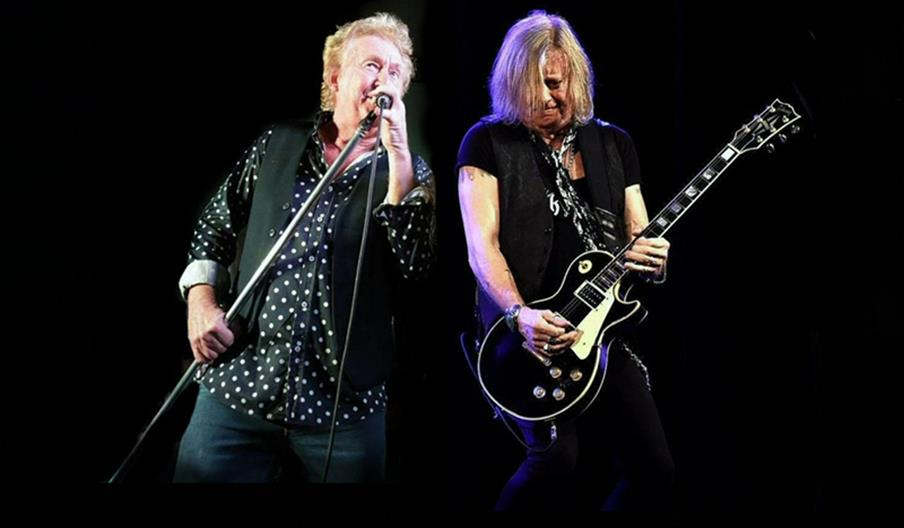 A man with blond hair sings into a microphone while wearing a black vest and polka-dot shirt, standing beside another man with long blond hair playing