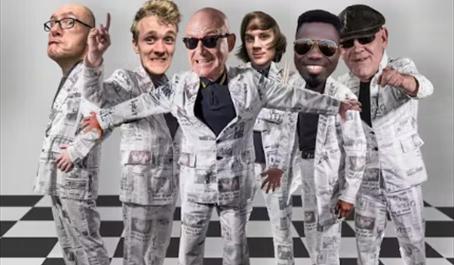 Five men wearing matching white suits with newspaper print stand on a black-and-white checkered floor, posing energetically. Above them, large red tex