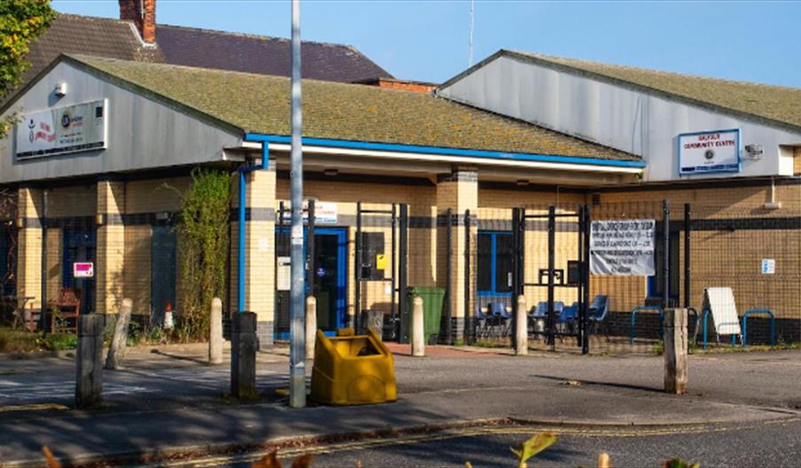 Single-story Balfour Community Center building with yellow brick walls, blue trim, and signs by the entrance, enclosed by a black metal fence; outdoor