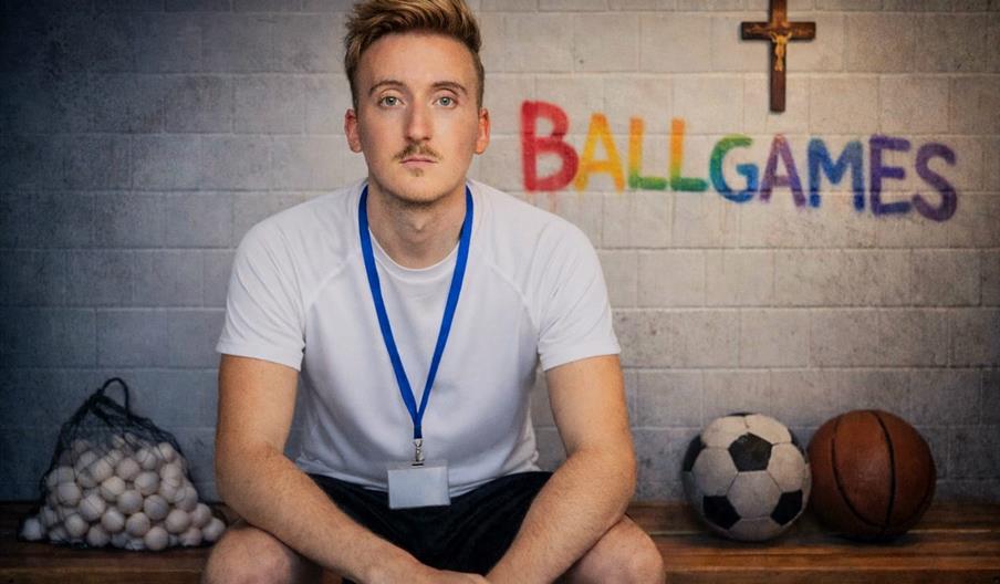 A young man with light hair, wearing a white athletic shirt and a blue lanyard, sits on a bench in a locker room. Behind him are a mesh bag full of ba