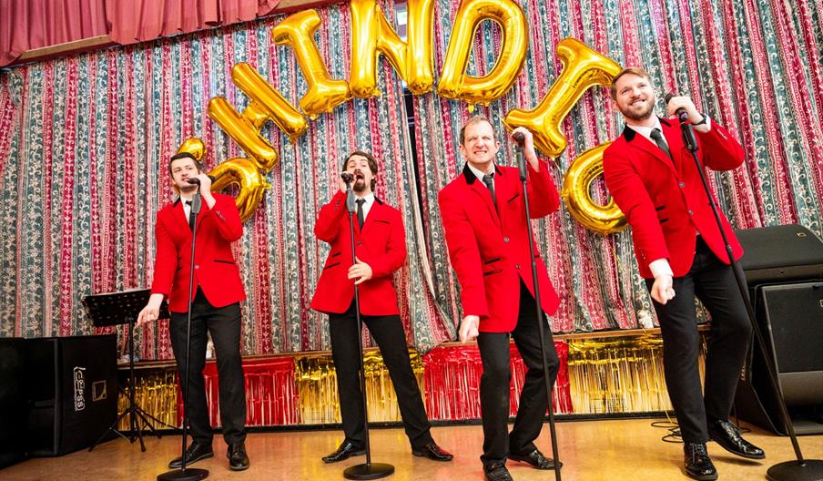 4 men in red jackets sing on a stage with the word 'SHINDIG' written in golden balloons behind them.
