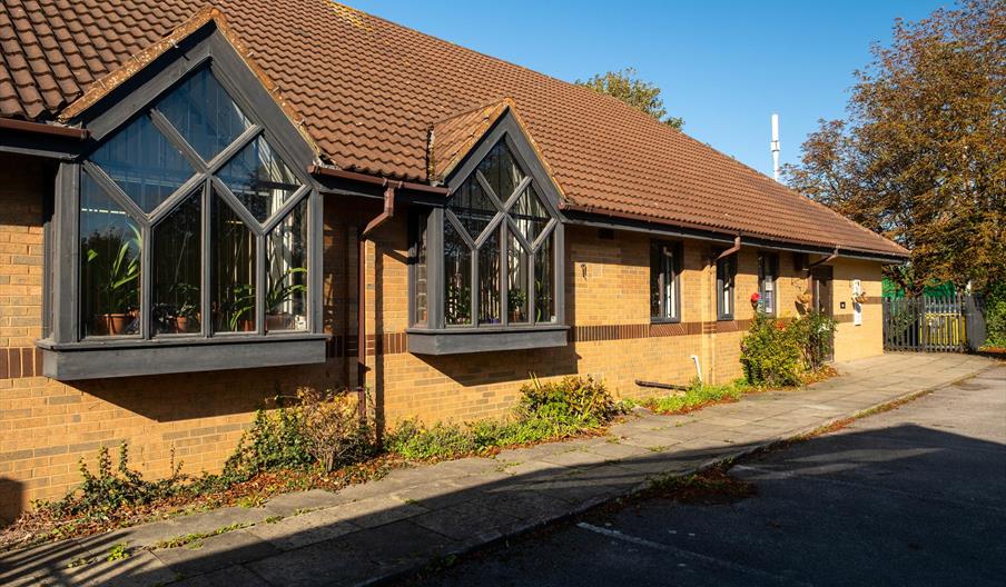 Single-story brick building with a brown tiled roof, featuring two large bay windows with black frames and decorative mullions, and some plants and sh
