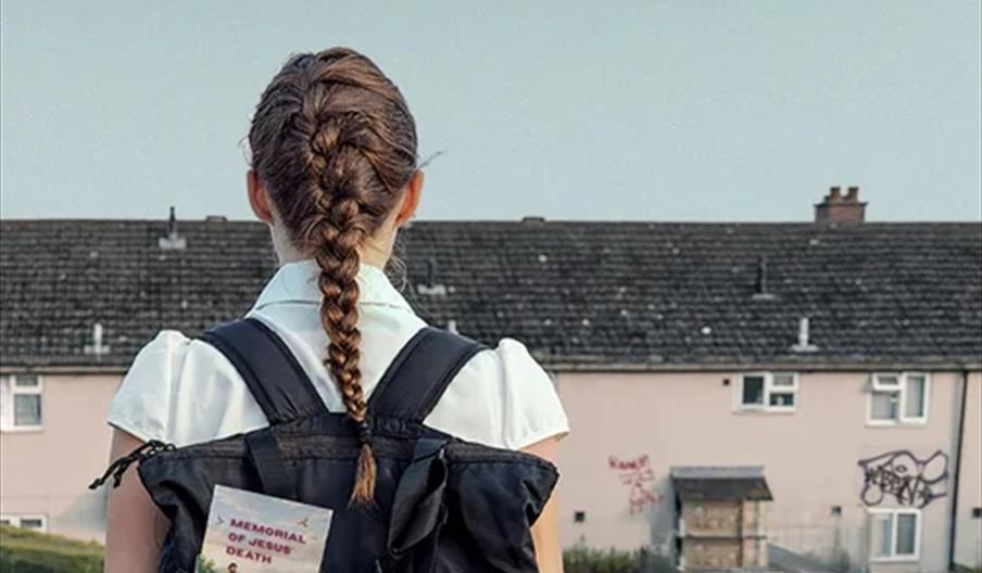 A girl with braided hair, seen from behind, stands outdoors wearing a backpack with a book titled "Memorial of Deaths" visible in a side pocket; subur