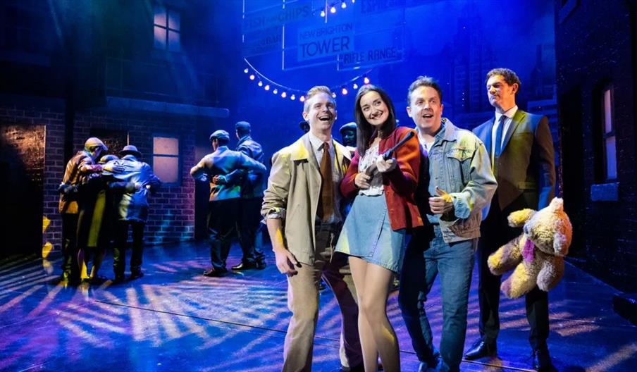 A brightly lit stage scene from a musical: several performers stand smiling in the foreground under blue and purple lights, dressed in mid-20th-centur