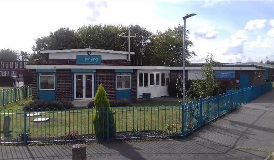 A single-story brick building with a sign reading "playing" above the entrance, surrounded by a blue metal fence and a small, neatly kept garden. Ther