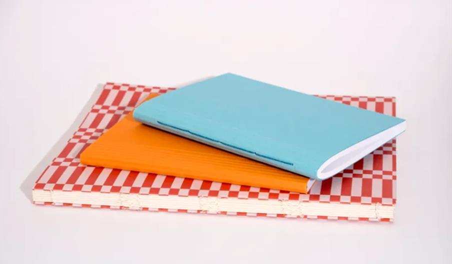 Three notebooks are stacked on a white surface: a blue notebook on top, an orange notebook in the middle, and a larger red-and-white checkered noteboo