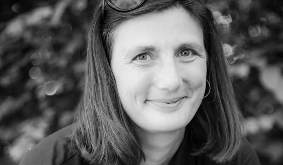 Black and white photo of a woman with shoulder-length hair, smiling softly at the camera, wearing sunglasses on her head and a hoop earring, with a bl