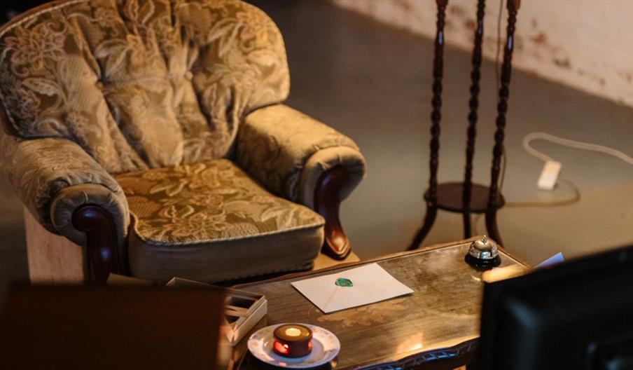 A vintage armchair with a floral pattern sits next to a glass-top coffee table holding a sealed envelope, a bell, and a lit candle in a small dish.