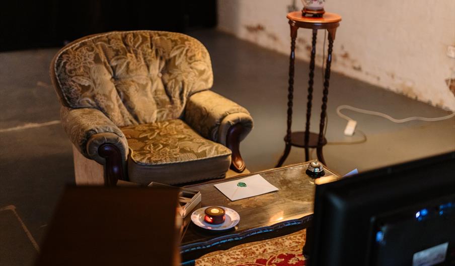 A vintage upholstered armchair sits next to a small wooden table holding a floral lamp; in front of the chair is a glass coffee table with a sealed en