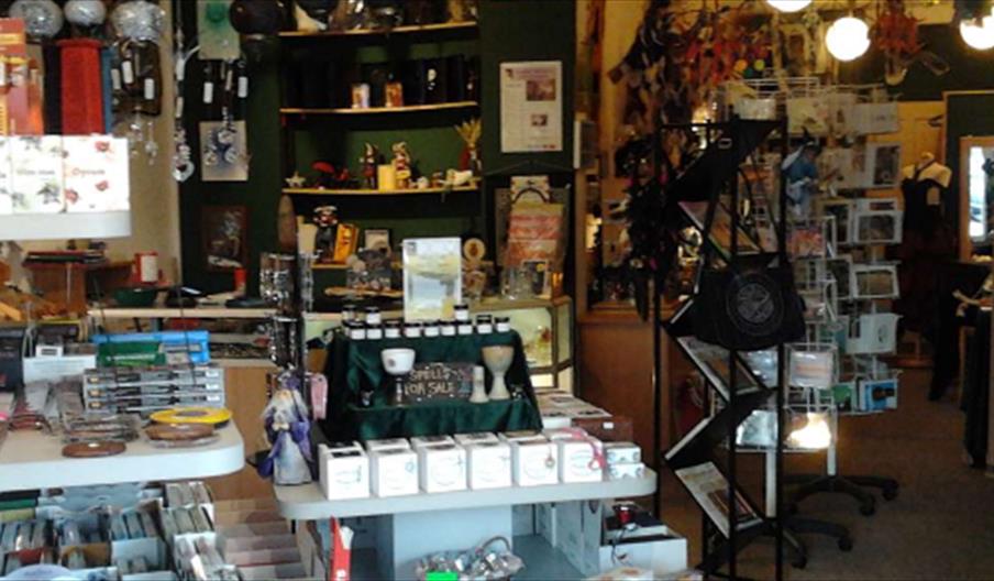 A small shop interior, Broomsticks with shelves and displays filled with various goods, including books, cards, candles, figurines, and metaphysical o