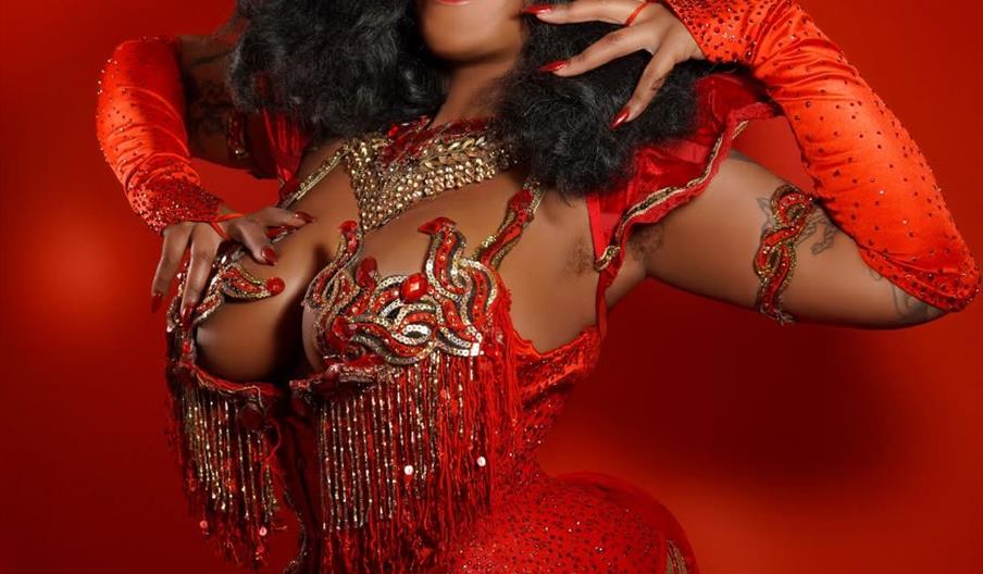 A performer in a sparkling red and gold burlesque costume with dramatic makeup and black curly hair poses confidently against a red background. Event