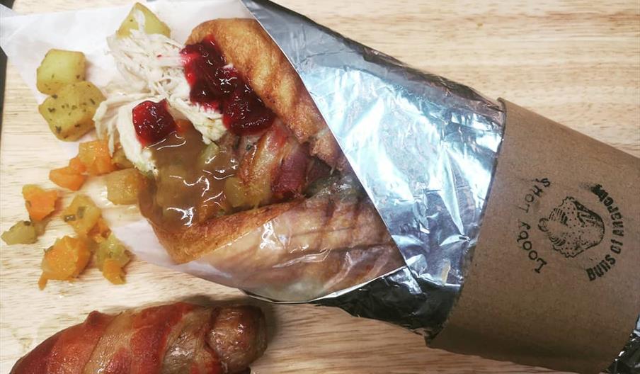 A handheld sandwich wrapped in foil and paper, filled with turkey, stuffing, cranberry sauce, and gravy, sits on a wooden board with a side of diced r